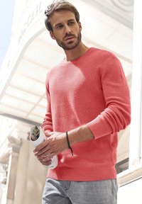 Koralfarbener Pullover mit strukturiertem Muster, runder Ausschnitt, gerippte Bündchen und Saum; graue Hose; hält eine gerollte Zeitschrift; Hintergrund im Freien.