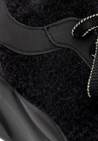 Zapatilla negra con una parte superior texturizada y fuzzy, y suela de goma negra lisa. Cuenta con un acento negro mate y cordones a rayas en contraste.