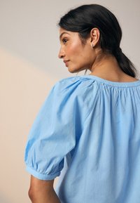 Hellblaues Baumwolloberteil mit Puffärmeln und gerafftem Halsausschnitt. Strukturierter Stoff mit kleinen, erhabenen Punkten überall.