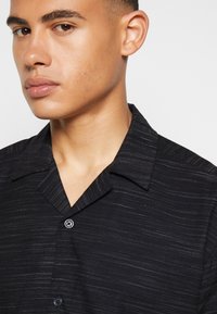 Black short-sleeve shirt with a textured pattern, featuring a notch collar and a button closure at the front.