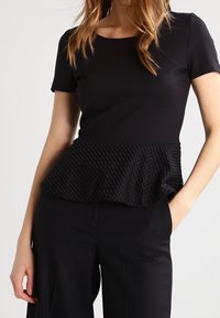 Černý top s krátkým rukávem, který má texturovaný peplum lem s vzorovaným, mírně volánkovým designem, spárovaný s černými kalhotami s širokými nohavicemi.