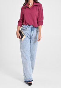 Femme portant un chemisier magenta à boutons avec des manches retroussées, un jean taille haute bleu clair, des talons noirs, et tenant une petite pochette rectangulaire.