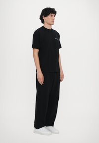 Svart kortermet t-skjorte, løs passform, rund hals, med subtil merkevare. Kombinert med svarte bukser og hvite sneakers, minimal design.
