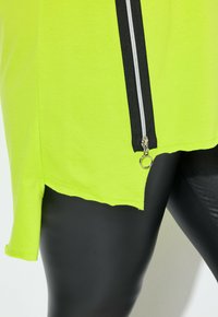 Camisa verde neón con cierre lateral, detalles en negro, dobladillo con aberturas y tejido suave y ligero; combinada con leggings negros.