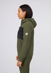 Olive green fleece hoodie with a black panel. Features a front zipper, two side pockets, and a bear logo on the left sleeve.