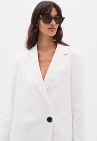Bela lanena blazer s klapnom, zapenjanjem na en črn gumb in dvema sprednjima žepoma; oblečen z očali za sonce s tortoiseshell vzorcem in ostrimi robovi.