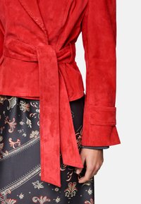 Red suede wrap jacket with long sleeves, tie waist detail, and textured finish, paired with a patterned skirt featuring floral motifs.
