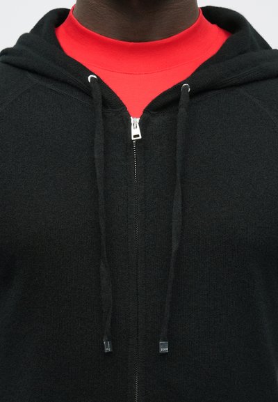 Pessoa vestindo uma camiseta vermelha de gola redonda debaixo de um casaco preto com zíper e cordões, mostrada do pescoço até o meio do peito.