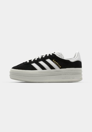 Black suede sneaker with white leather accents, three white stripes, flat laces, and a thick grey platform sole. Gold "Gazelle" branding.