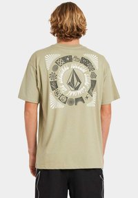 Vista trasera de una camiseta verde oliva claro con un gran gráfico circular que presenta varios patrones y el texto "THE ORIGINAL VOLCOM STONE COMPANY."
