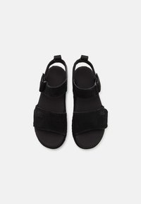 Zwarte suede sandalen met twee brede banden, verstelbare enkelriem, platte zool en UGG-logo aan de zijkant. Zachte textuur en minimalistisch ontwerp.