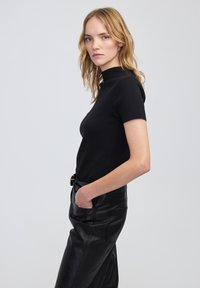 Kobieta z falującymi blond włosami, ubrana w dopasowaną czarną bluzkę z krótkim rękawem oraz czarne skórzane spodnie z wysokim stanem, trzymająca jedną rękę w kieszeni.