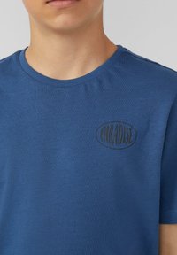 Blauwe T-shirt met korte mouwen, gemaakt van zachte stof, met een ronde hals en een zwart "PARADISE" logo in een ovale vorm op de linkerborst.