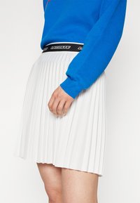 Jupe plissée blanche avec une taille noire portant la marque "LACOSTE". Un haut ajusté bleu avec un col rond et des manches longues.