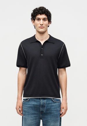 Jeune homme aux cheveux bouclés foncés portant un polo noir à manches courtes avec des liserés blancs et un jean bleu, debout face à l’avant.