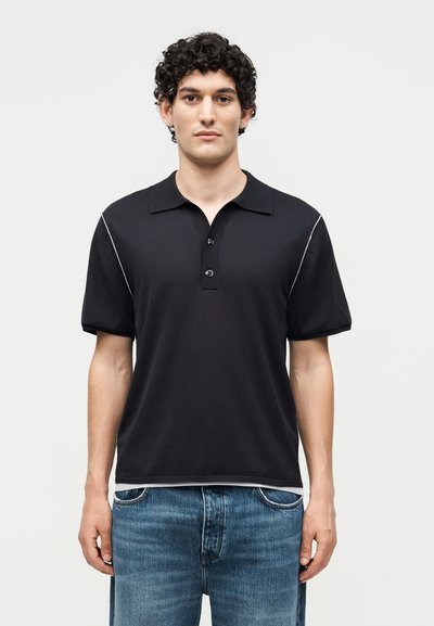 Jeune homme aux cheveux bouclés foncés portant un polo noir à manches courtes avec des liserés blancs et un jean bleu, debout face à l’avant.