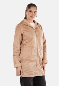 Cappotto in finta pelle scamosciata beige con cappuccio, zip frontale, due grandi tasche e polsini in morbido pile. Esterno testurizzato con una finitura liscia.