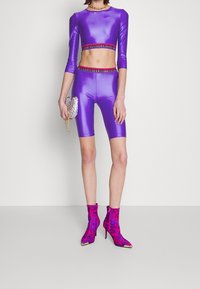 Purple cropped top and matching bike shorts with shiny texture; embellished ankle boots in pink with black logo pattern; silver clutch.