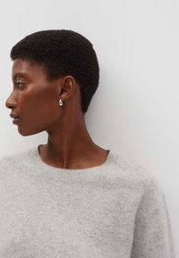 Femme aux cheveux bouclés courts, portant un pull gris, de profil à gauche, avec de petites boucles d'oreilles en argent en forme de goutte contre un fond blanc uni.