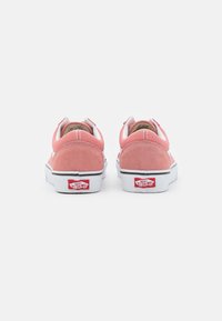 Różowe zamszowe sneakersy z białymi gumowymi podeszwami, mające czarny pasek, okrągły czubek i czerwone logo na pięcie.
