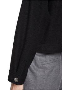 Černá strukturovaná bunda s dlouhým rukávem a knoflíkovými manžetami, doplněná o šedé kalhoty na míru s kapsou na boku.