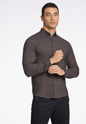 Donkerbruine blouse met lange mouwen en knopen, gemaakt van een gestructureerde stof. Heeft een klassieke kraag en zwarte knopen, gecombineerd met zwarte broeken.