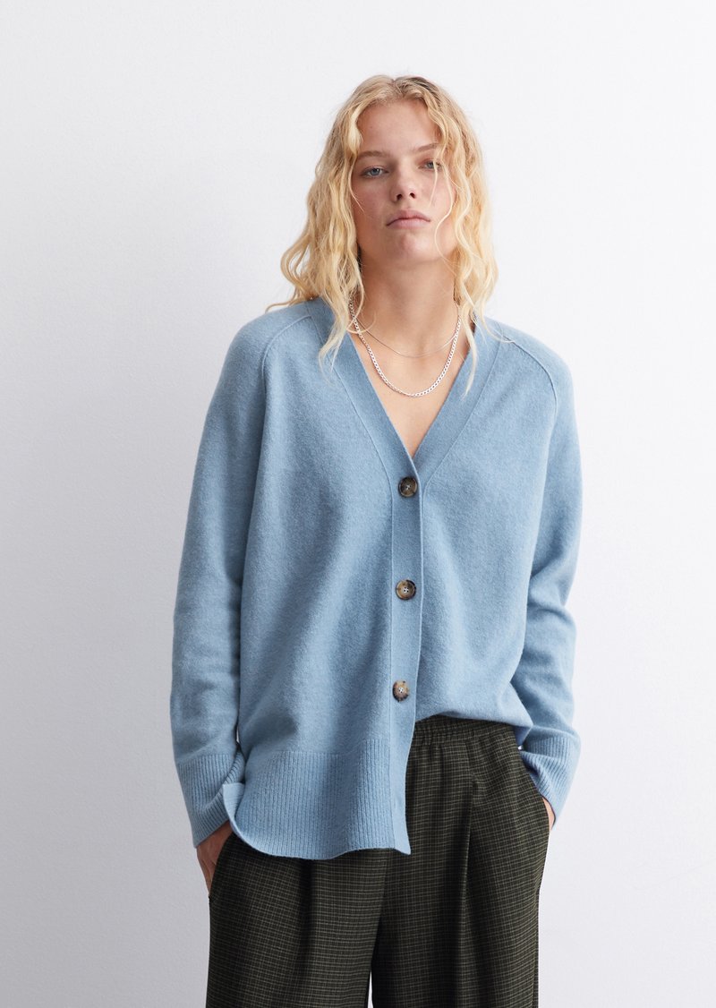 Helles blaues Wollcardigan mit V-Ausschnitt, fünf Knöpfen in der Mitte, gerippten Bündchen und lockerer Passform, kombiniert mit dunklen karierten Hosen.