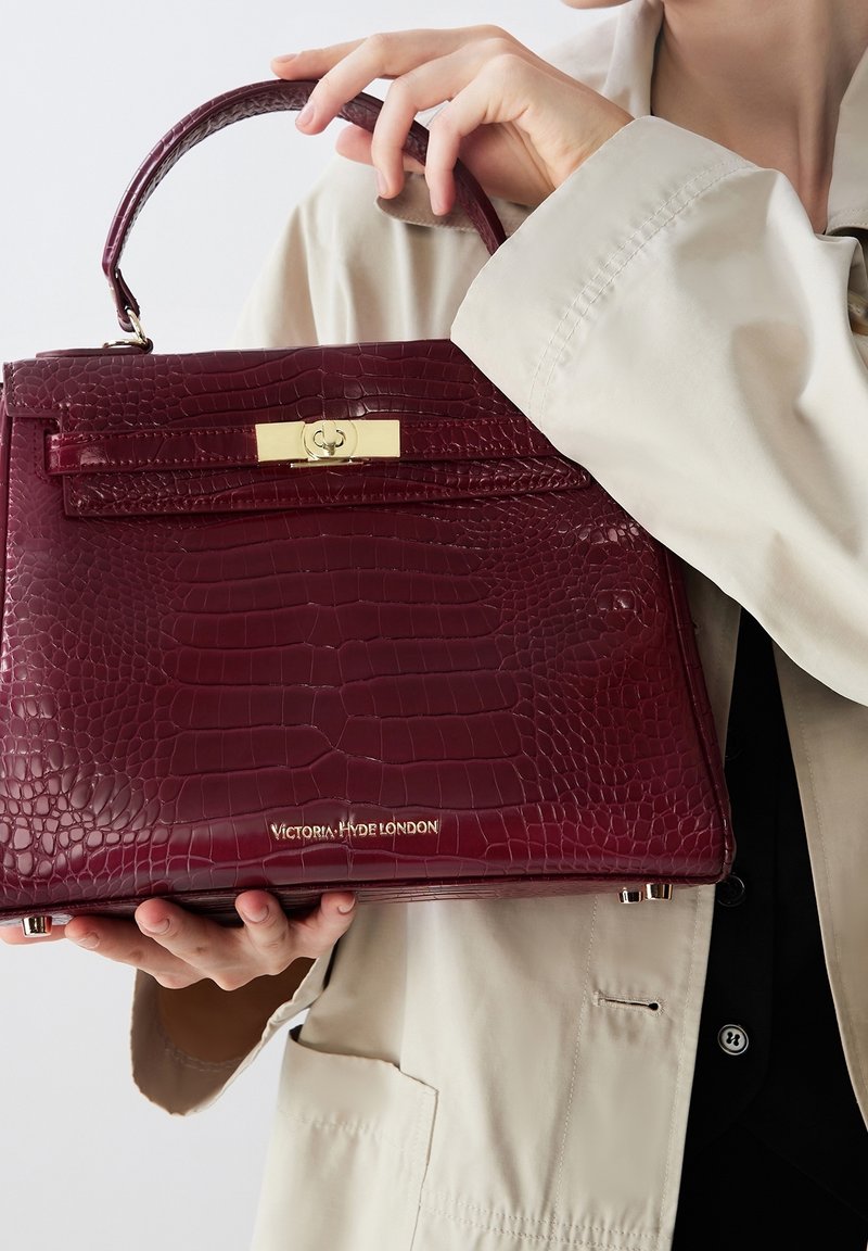 Burgundy toteväska med präglad krokodilstruktur, guldfärgad h årdvara och ett handtag. Har en logotyp i basen och en strukturerad form.