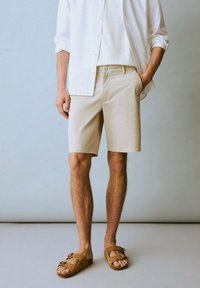 Shorts beige en coton avec une coupe ajustée, associés à une chemise blanche. Le modèle porte des sandales marron avec des accents de boucle.