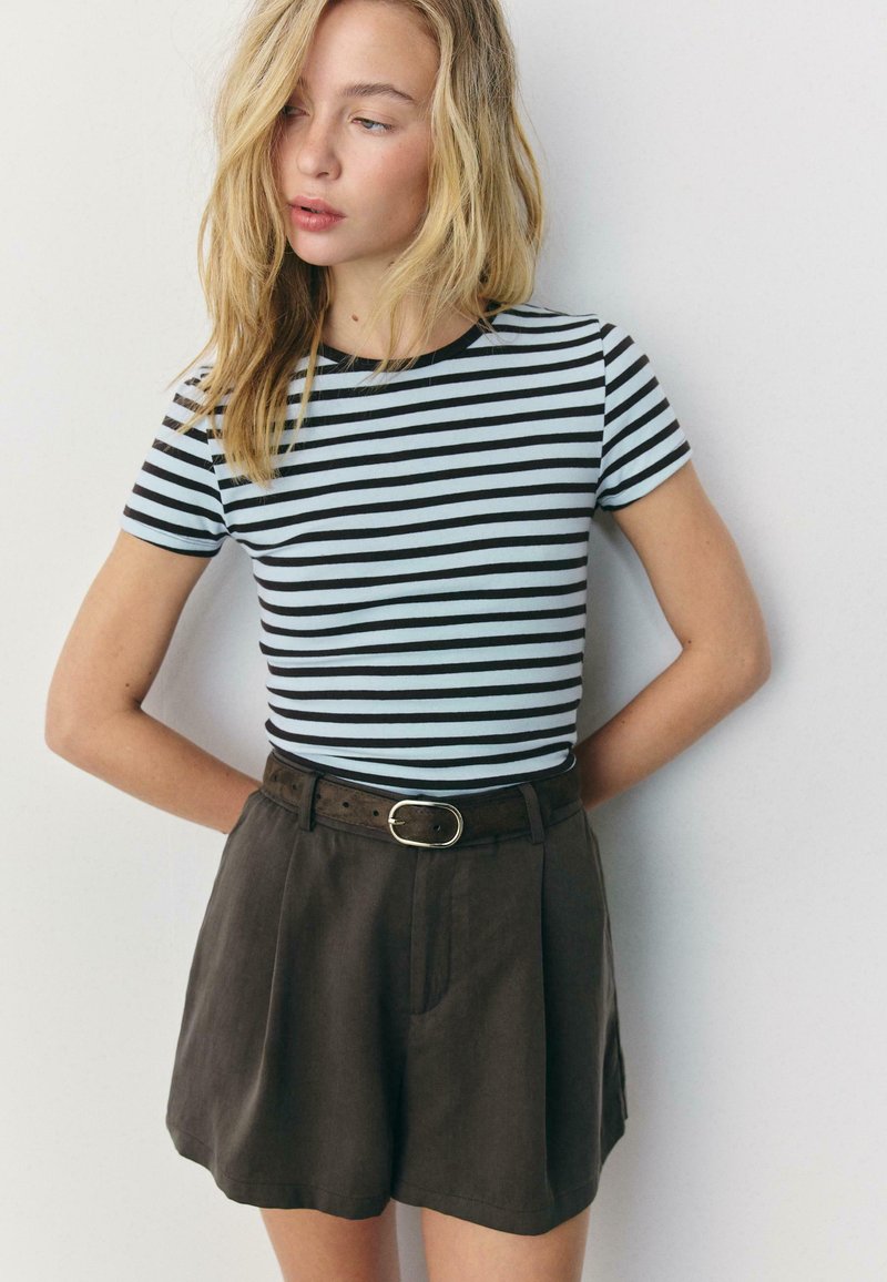 Ung kvinde med blondt hår iført en kortærmet sort og hvid stribet skjorte, der er stoppet ned i højtaljede mørke shorts med bælte.