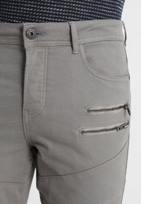Graue Baumwollhose mit Slim-Fit, ausgestattet mit zwei Reißverschlusstaschen vorne und einer dezenten strukturierten Oberfläche. Metallknopfverschluss.