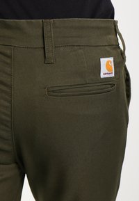 Olijfgroene broek van stevige stof, met een achterzak en een vierkant, geborduurd Carhartt-logo in oranje en wit.