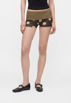Person iført sort og gulstribet taljebælte, sorte shorts med store gule polkaprikker, sorte Mary Jane-sko og en sort top.