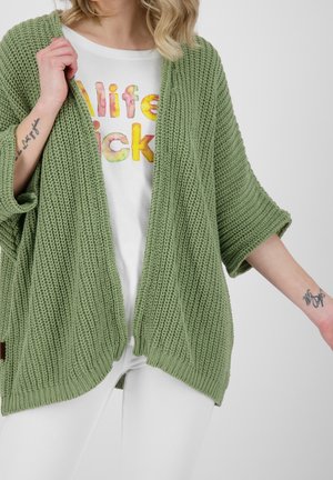 Femme portant un cardigan en laine vert clair par-dessus un t-shirt blanc avec le texte coloré « life kicks », bras légèrement étendus montrant des tatouages aux poignets.