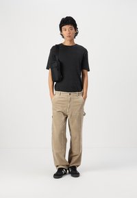 Czarny t-shirt z krótkim rękawem, beżowe spodnie z koralików z bocznymi kieszeniami, czarna torba na ramię, czarne trampki. Jednolity tło.