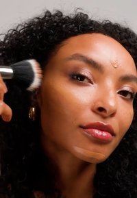 Vrouw met krullend haar brengt foundation aan op wang en kin met een make-upkwast tegen een neutrale achtergrond.