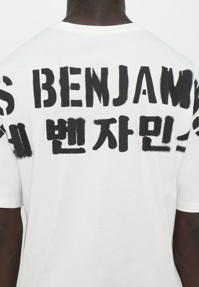 T-shirt blanc avec un grand texte noir au dos. Le tissu semble doux avec une coupe décontractée et des manches courtes.