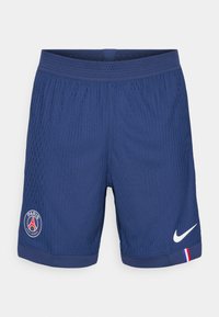 Tums zila sporta bikses no teksturēta auduma, ar Nike logo, Parīzes emblēmu un sarkano-zilo-balto akcentu uz apakšmalas.