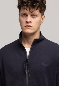 Granatowa kurtka na zamek z fakturowanej tkaniny, z podniesionym kołnierzem, kontrastowym zamkiem z uchwytem oraz małym logo na lewej piersi.