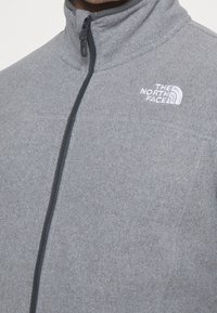Gray fleece jacket with a high collar, front zip closure, and a white logo on the left chest. Features smooth texture and contrast stitching.