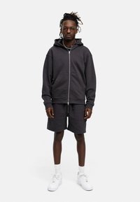 Dunkelgrauer Reißverschluss-Hoodie mit einer Vordertasche, kombiniert mit passenden Shorts. Getragen mit weißen Sneakers und Knöchelsocken. Einfaches, lässiges Design.