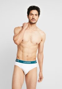 Fehér pamut férfi alsónadrág teal színű derékkal, amelyen a "LACOSTE" felirat található. Az anyag sima, a dizájn klasszikus és funkcionális.