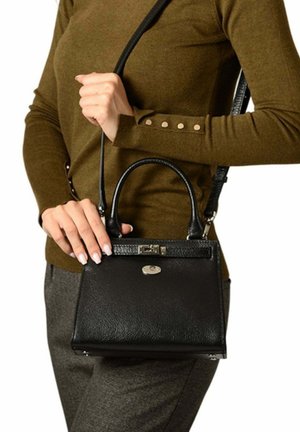 Femme portant un pull vert olive et un pantalon gris, tenant un sac à main en cuir noir avec des poignées supérieures et une bandoulière.