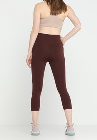 Kobieta w brązowych legginsach o wysokim stanie i beżowym biustonoszu sportowym, stojąca z jedną ręką na biodrze, zwrócona plecami.