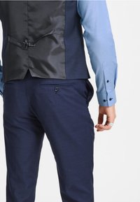 Pánský formální outfit s černou vestou s hladkou texturou a modrou košilí s manžetovými knoflíky, doplněný tmavě modrými kalhotami a decentním detailem na kapse.
