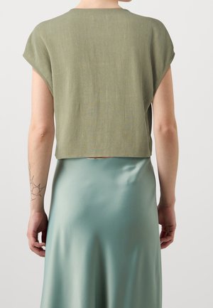 Person, die ein olivgrünes kurzärmeliges Oberteil und einen hellblauen Satinsrock trägt, von hinten gezeigt mit einem Tattoo am linken Unterarm.