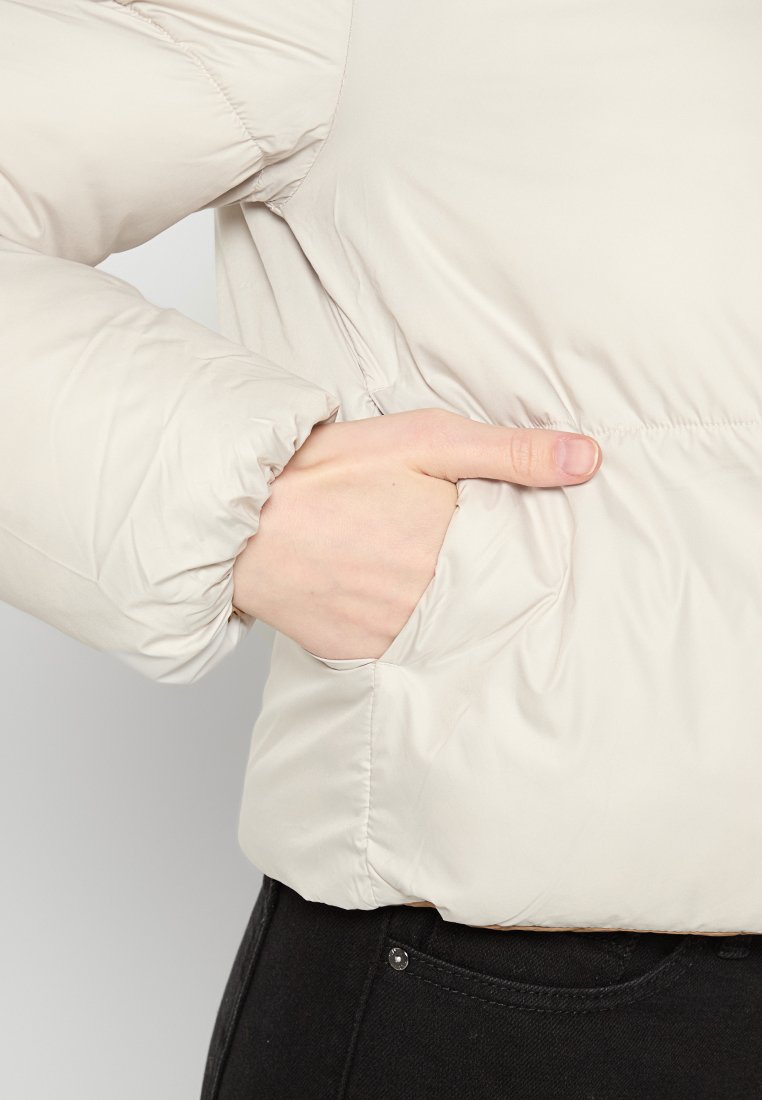Chaqueta acolchada de color beige claro con un bolsillo lateral visible, puños elásticos y una textura suave y ligeramente brillante. Detalles de costura a lo largo del cuerpo.