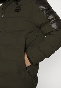 Chaqueta acolchada de color verde oscuro con detalles brillantes negros en los hombros, botones delanteros, bolsillos laterales y puños de punto para mayor calidez.