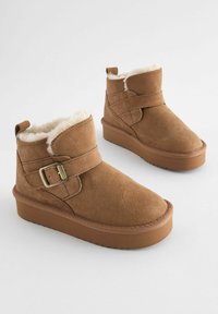 Stivaletti alla caviglia in suede beige con fodera in pelliccia sintetica, caratterizzati da una fascia larga e un dettaglio con fibbia. La suola in gomma elevata aggiunge altezza e aderenza.
