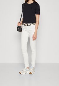 Top negro de manga corta con ribetes rojos y blancos, jeans blancos ajustados, cinturón negro, bolso pequeño negro y zapatillas gruesas blancas con detalles azules.